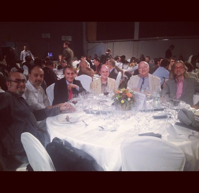 Siete hombres sentados a una mesa redonda con mantel blanco en la cena del Premio Planeta 2013; el de la derecha tiene el pelo largo y gafas