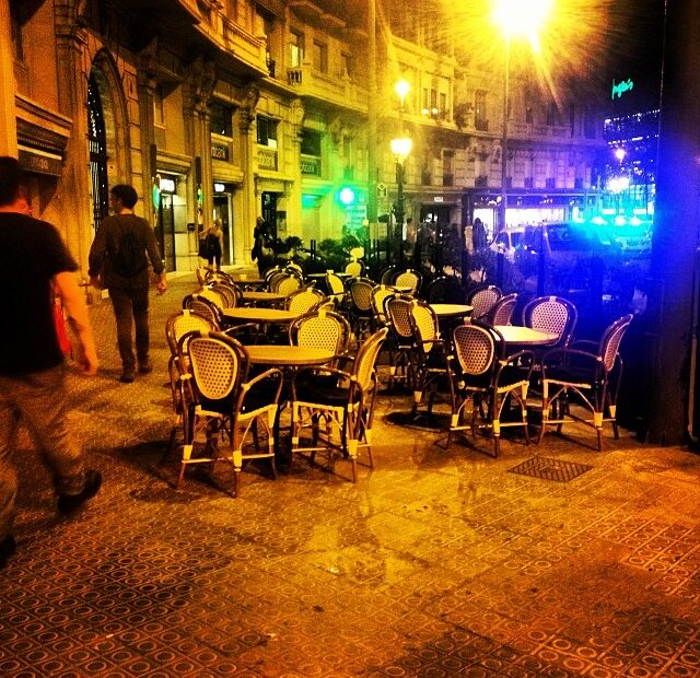 Terraza vacía de noche con sillas de rejilla y mesas redondas en una esquina iluminada de Barcelona
