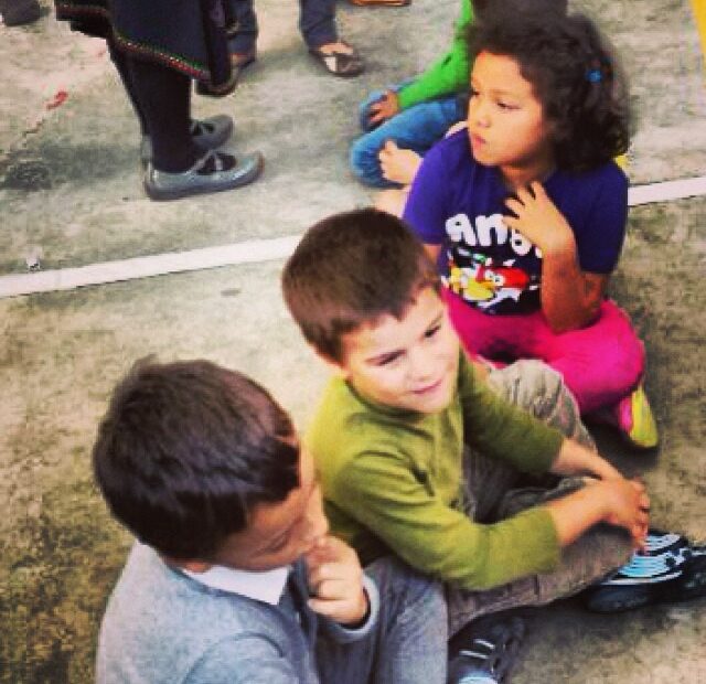 Tres niños sentados en el suelo de un patio, uno de ellos con camiseta verde, y otra niña con camiseta de Angry Birds detrás