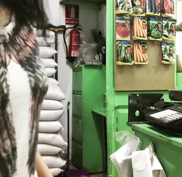 Una mujer en movimiento, de perfil, junto a sacos y un expositor de sobres de semillas en una tienda con muebles verdes