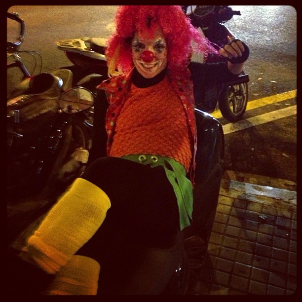 Payaso con peluca rosa sentado junto a unas motos en la calle