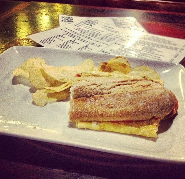 Bocadillo de tortilla francesa con patatas fritas sobre un plato blanco, con la carta de un bar al fondo