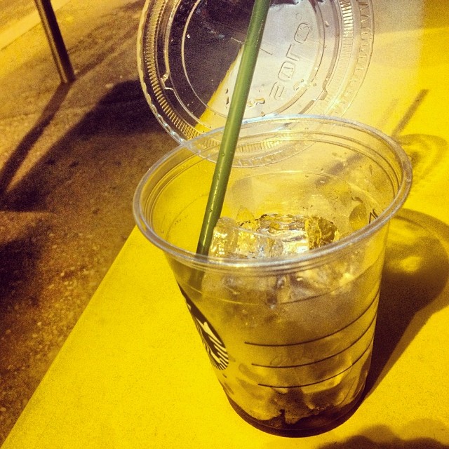Vaso de plástico con hielo y una pajita verde sobre un banco amarillo, de noche