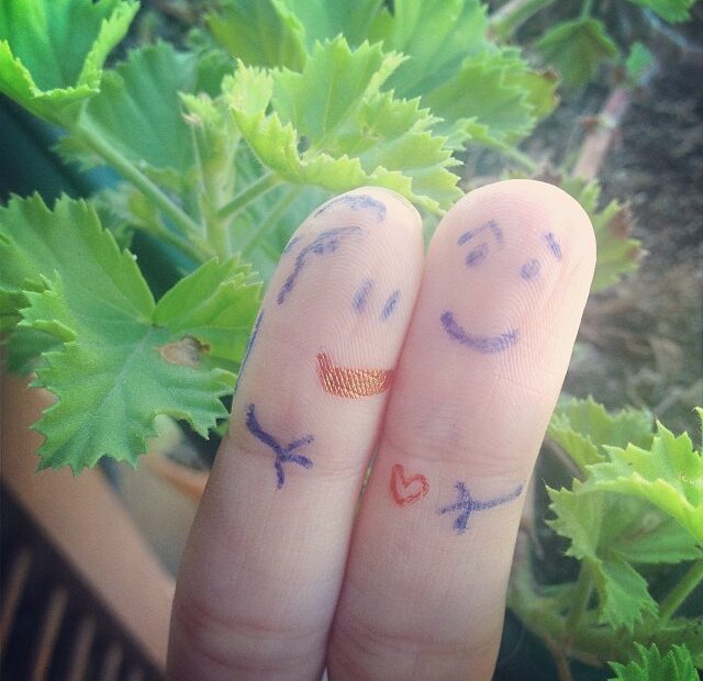 Dos dedos juntos con caras dibujadas a rotulador, uno con labios y el otro con un corazón, ante hojas de geranio