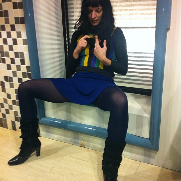 Roser Amills sentada en el alféizar de una ventana, con falda azul y medias oscuras, mirando una prenda que sostiene