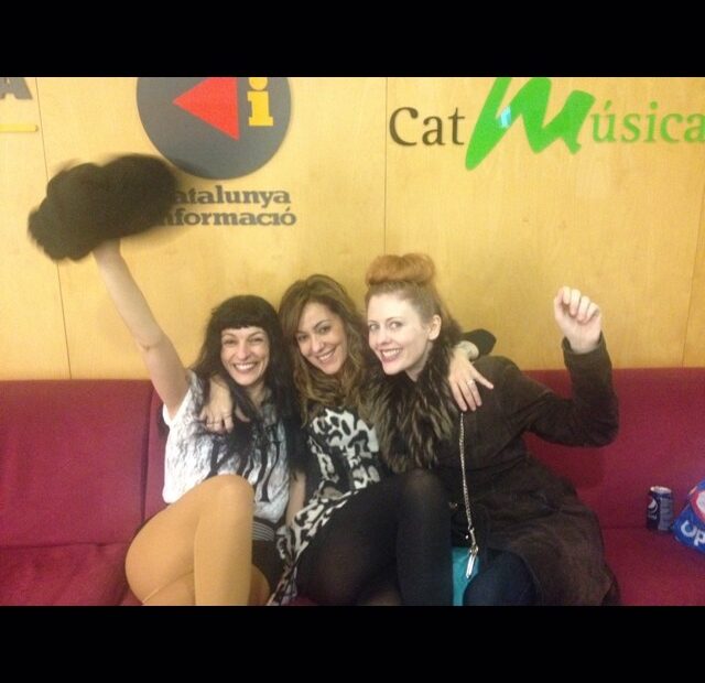 Roser Amills y dos mujeres abrazadas y sonrientes en un sofá rojo, con un pompón negro en alto, ante el cartel de CatMúsica