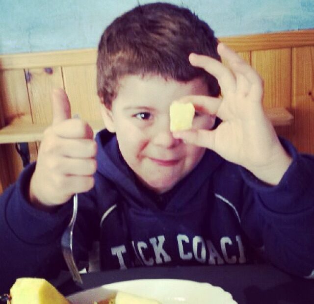 Niño con sudadera azul sonríe con un trozo de piña ante el ojo y el pulgar levantado