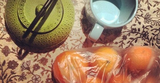 Tetera verde de hierro, taza y bolsa de mandarinas sobre un mantel estampado