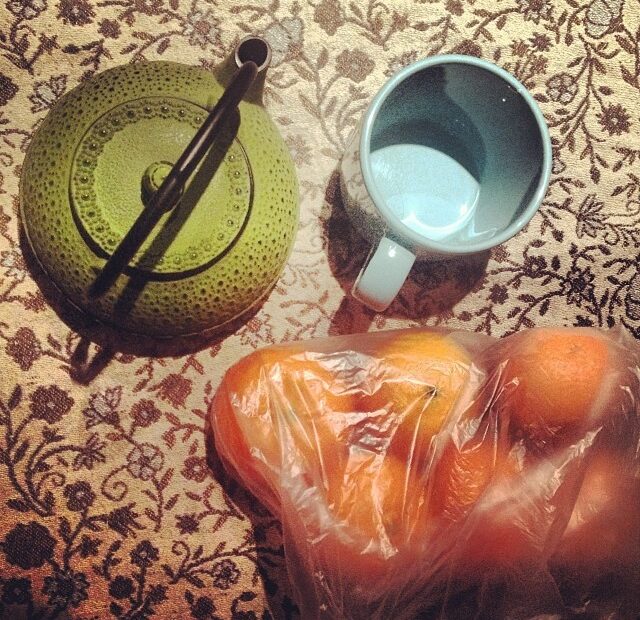 Tetera verde de hierro, taza y bolsa de mandarinas sobre un mantel estampado