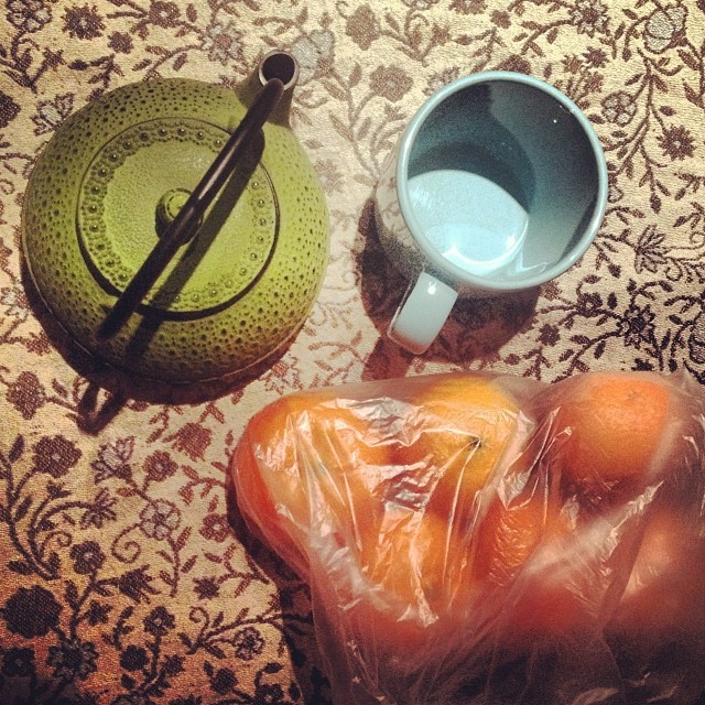Tetera verde de hierro, taza y bolsa de mandarinas sobre un mantel estampado