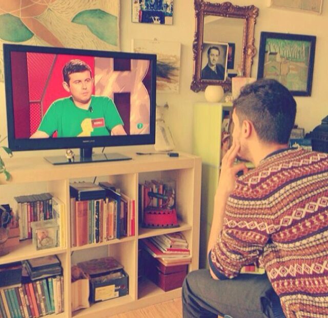Un jove assegut mira la televisió davant d'una prestatgeria plena de llibres