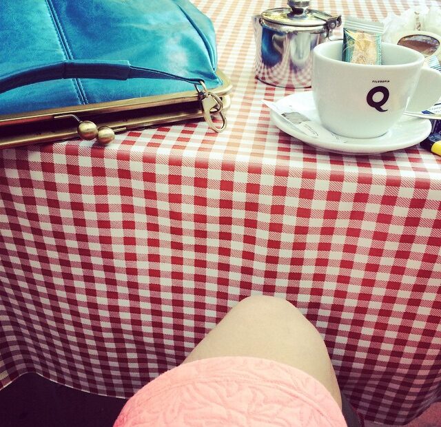Mesa de bar con mantel de cuadros rojos y blancos, una taza de café, un bolso azul y el borde de una falda rosa