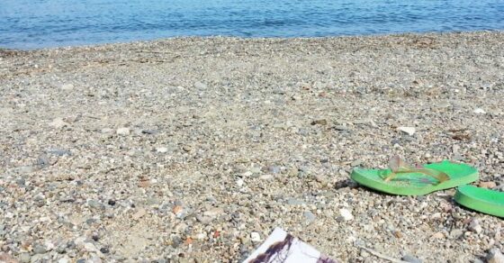 Ejemplar de «Sé buena» sobre una playa de guijarros, junto a unas chancletas verdes, con el mar al fondo
