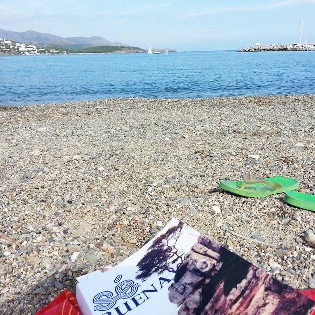 Ejemplar de «Sé buena» sobre una playa de guijarros, junto a unas chancletas verdes, con el mar al fondo