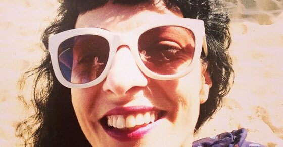Selfi d'una dona somrient amb ulleres de sol blanques i el cabell arrissat, amb la sorra de fons