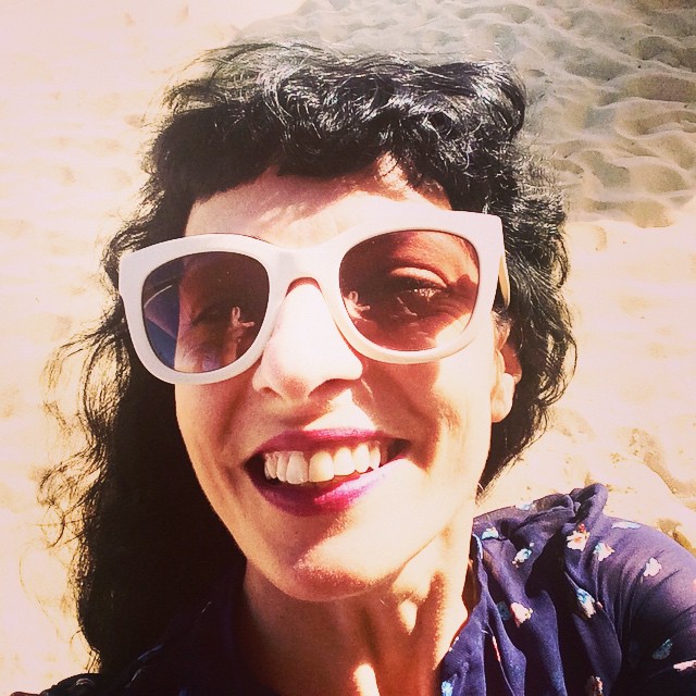 Selfi d'una dona somrient amb ulleres de sol blanques i el cabell arrissat, amb la sorra de fons