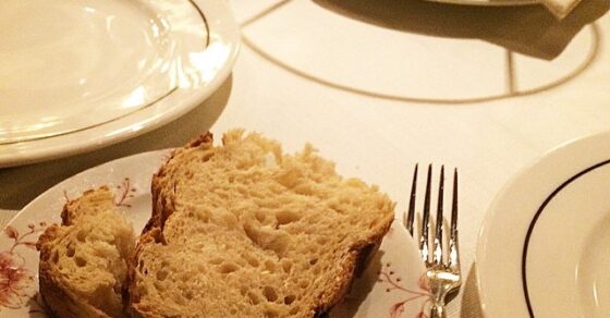 Un trozo de pan sobre un plato de porcelana decorado, con cubiertos y una copa en la mesa de un restaurante