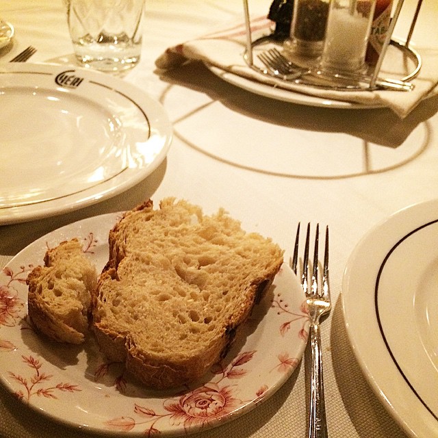 Un trozo de pan sobre un plato de porcelana decorado, con cubiertos y una copa en la mesa de un restaurante