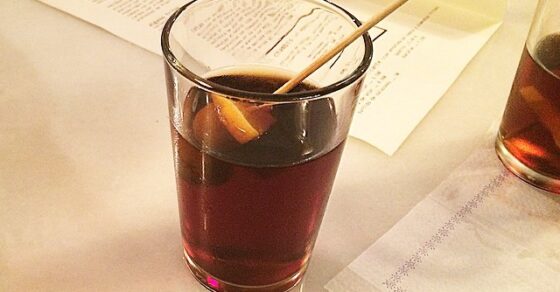 Vaso de vermut con una rodaja de naranja y un palillo sobre la barra de mármol de La Cava, con la carta de papel al fondo