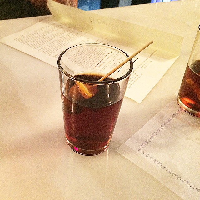 Vaso de vermut con una rodaja de naranja y un palillo sobre la barra de mármol de La Cava, con la carta de papel al fondo