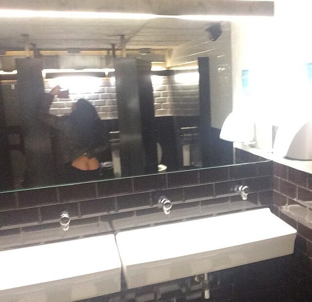 Lavabo con una pica corrida de porcelana blanca y un espejo largo y alto, en el que se refleja una persona haciendo la foto