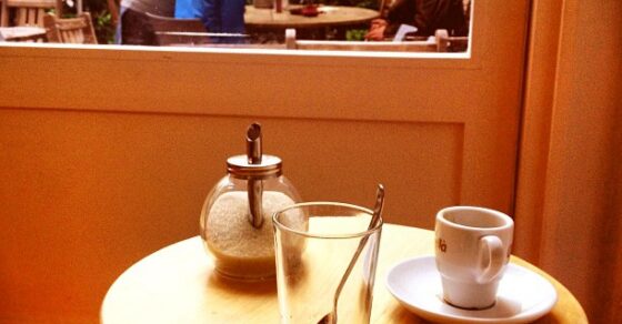 Mesa redonda de madera con una taza de café, un vaso con una cucharilla y un azucarero, ante una ventana que da a un patio con gente