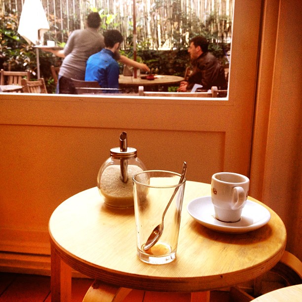Mesa redonda de madera con una taza de café, un vaso con una cucharilla y un azucarero, ante una ventana que da a un patio con gente