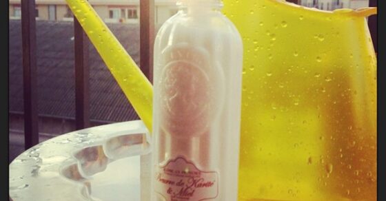 Botella de leche corporal Jeanne en Provence de karité y miel junto a una regadera amarilla en un balcón