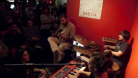 Músicos tocando en un pequeño local de paredes rojas con un cartel de «Poesia», ante el público