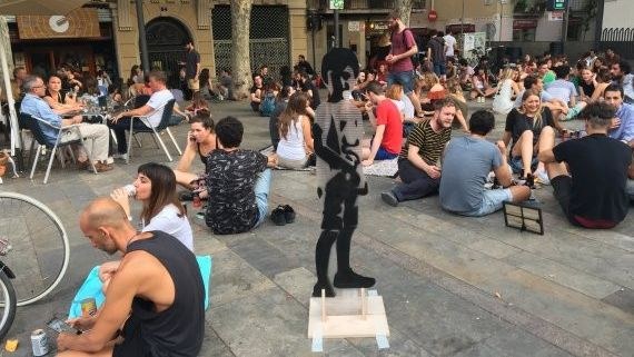 Una plaza de Barcelona llena de gente sentada en el suelo y en las terrazas, con una pequeña escultura negra en primer plano