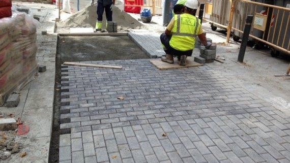 Un obrero con chaleco amarillo coloca adoquines grises en una acera en obras, junto a unas vallas