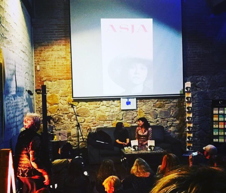Roser Amills y M. Àngels Jover conversan sentadas ante el público, con la cubierta de Asja proyectada en la pantalla