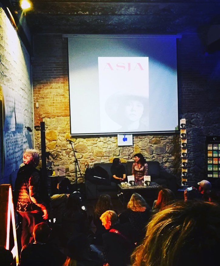 Roser Amills y M. Àngels Jover conversan sentadas ante el público, con la cubierta de Asja proyectada en la pantalla
