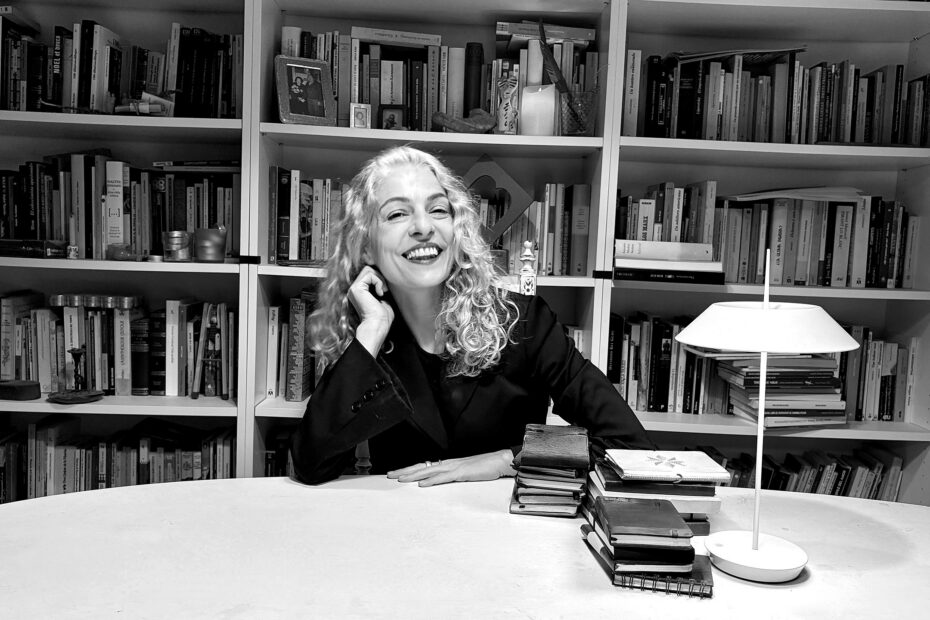 Roser Amills, con chaqueta oscura, sonríe apoyando la mano en la mejilla ante su biblioteca. Fotografía en blanco y negro.