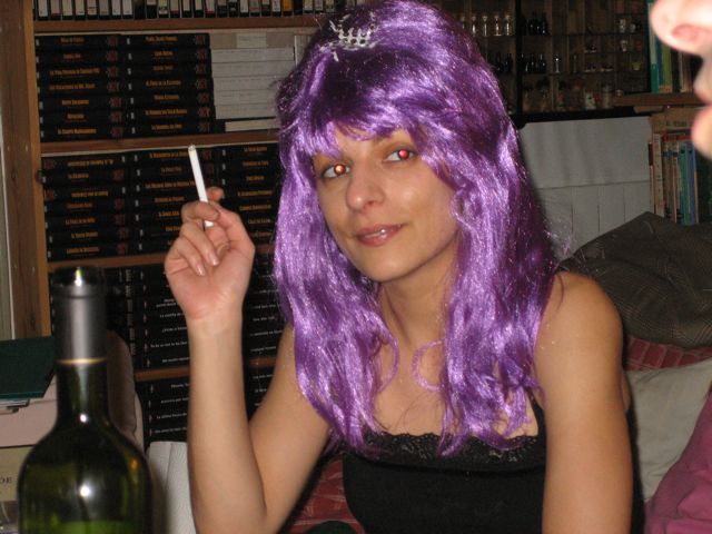 Roser Amills, con peluca lila y un cigarrillo, sentada ante una estantería de libros