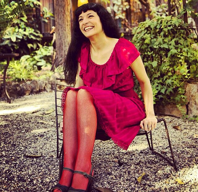 Roser Amills sonríe sentada en una silla de jardín, con vestido rojo y medias de rejilla; foto de Guy Aelbrecht