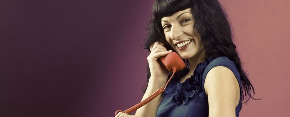 Roser Amills sonriente, con vestido azul, hablando por un teléfono rojo ante un fondo granate