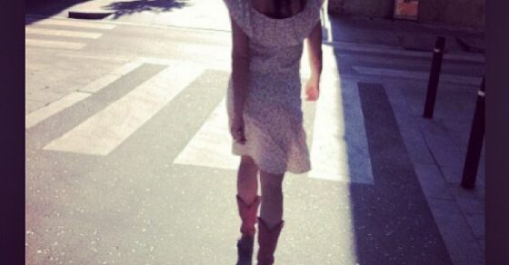 Roser Amills de espaldas, con vestido claro y botas rosas, cruzando una calle soleada