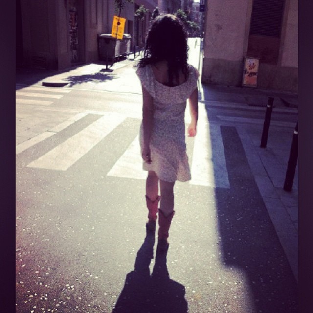 Roser Amills de espaldas, con vestido claro y botas rosas, cruzando una calle soleada
